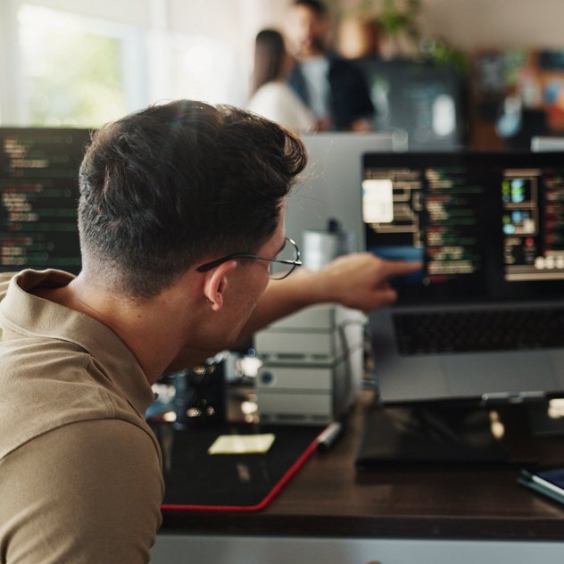 Prozessautomationssoftware im S88-Umfeld  Zwei Entwickler arbeiten an Codescreens am Schreibtisch; einer zeigt dem anderen etwas auf dem Laptop, im Büro mit Monitoren.